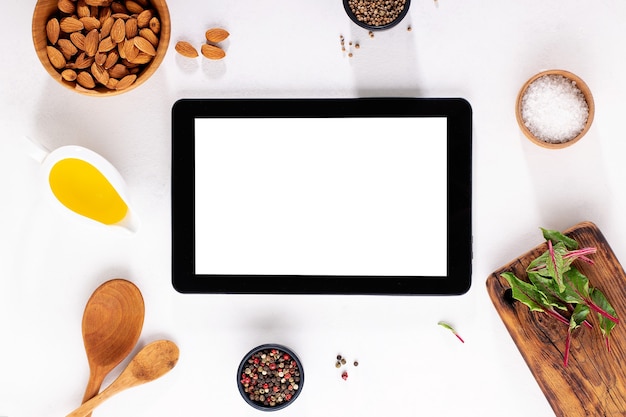 Digitale tablet met leeg scherm op witte tafel met kruiden, olie, bord, zout en noten.