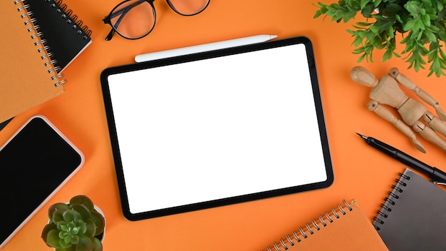 Digitale tablet met leeg scherm, smartphone, kamerplant, bril en notitieboekje oranje achtergrond.