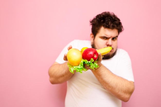 dikke jonge man in wit t-shirt staat op roze achtergrond en houdt fruit en groenten in zijn hand