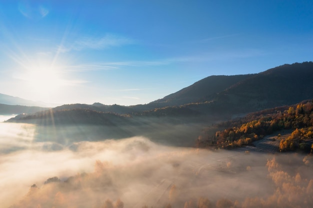 Dikke laag mist die hoge bergen bedekt met terracotta en groene bomen die groeien onder een helderblauw, wolkenloos luchtpanorama