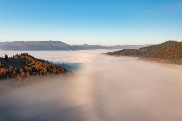 Dikke laag mist die regenbooghoge bergen bedekt met terracotta en groene bomen die groeien onder een helderblauw, wolkenloos luchtpanorama