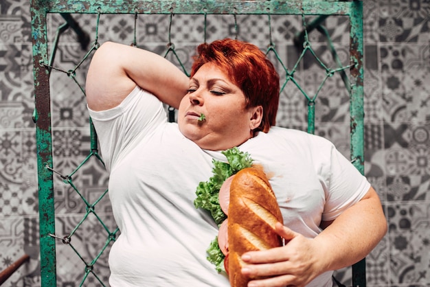 Dikke vrouw met sandwich in handen liying op het bed, bulimisch en overgewicht. Ongezonde levensstijl, zwaarlijvigheid