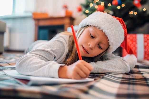 Diligent girl is lying on blenket on floor and writing letter to Santa. She uses red pencil. Girl is serious and concentrated. She is in room alone. There are Christmas tree with presents behind her.