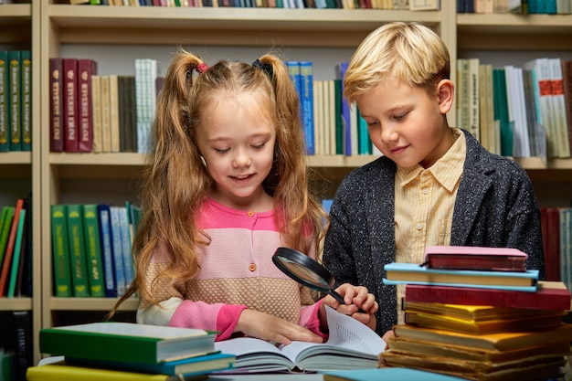 図書館に本を持っている勤勉な生徒、男の子と女の子はより良い学習と読書のために虫眼鏡を使用しています