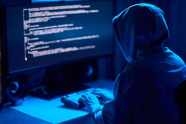暗く照らされた部屋で仮面をかぶった人物が謎のハッカーを暗示するコンピュータにコードをタイプします