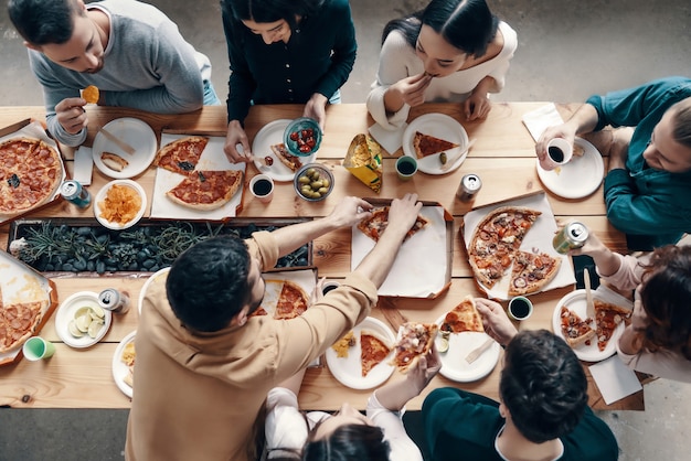 Diner onder vrienden. Bovenaanzicht van jonge mensen in vrijetijdskleding die pizza's plukken tijdens een etentje binnenshuis