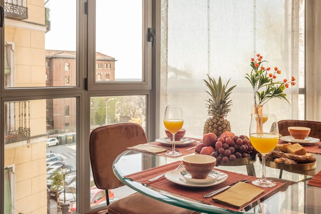 Dining room with a circular glass table full of assorted fruits glasses of orange juice and brown chairs next to a bay window with a view