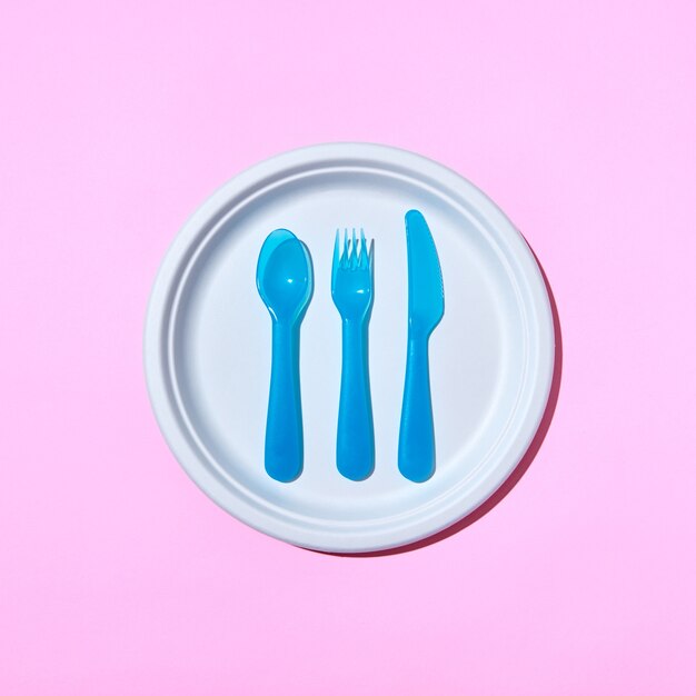 Dining set of plastic cutlery on a white plate on a pink pastel background with copy space. Flat lay.