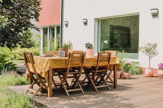 Dining table covered with orange tablecloth standing on wooden terrace in green garden