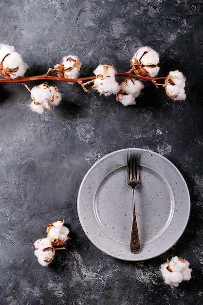 写真 夕食の場所の設定。暗い質感の表面に綿の花で飾られた銀のフォークが付いた青いモダンなセラミックプレート。上面図、フラットレイ。