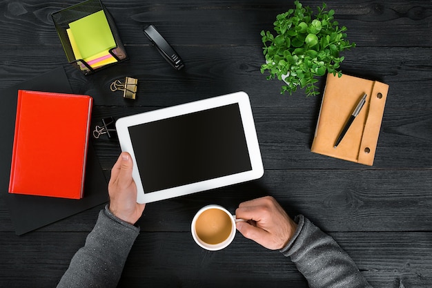 Direct boven weergave van menselijke handen met digitale tablet. Digitale tablet, agenda, koffiekopje en potplant op bureau. Man aan het werk vanuit huis.