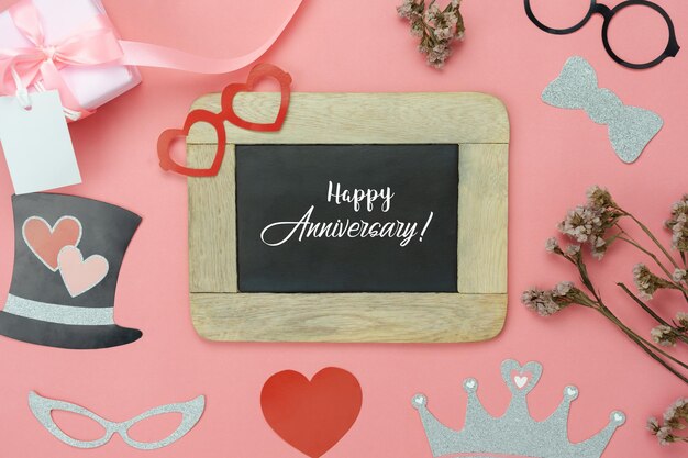 Directly above shot of blackboard with happy anniversary message and decorations over coral background