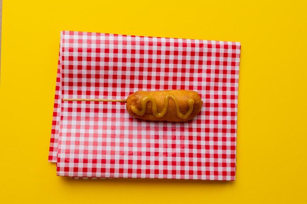 Directly above shot of corn dog with mustard sauce on checked pattern napkin over yellow background. unaltered, unhealthy food, meat, sausage and fried food