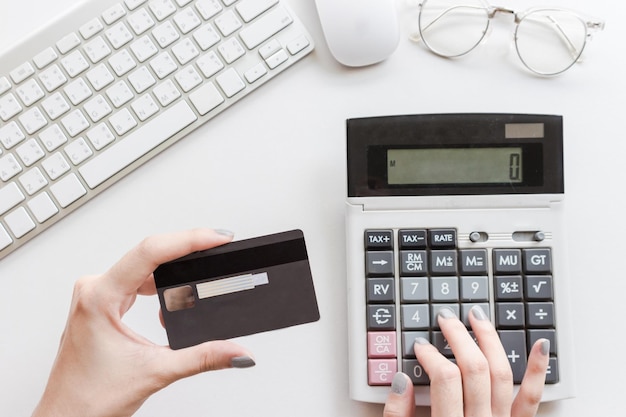 Directly above shot of woman using calculator by keyboard and mouse while holding credit card over white background