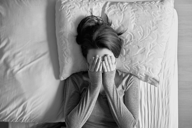 directly above shot of young woman covering face with hands while lying on bed at home