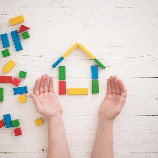 Directly above on wooden colorful bricks on a white wooden backgroundHands of a man protect a house