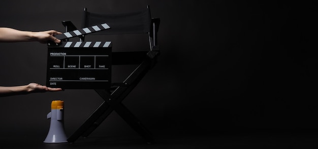 Director chair and magaphone and hand is hold clapper board on\
black blackground