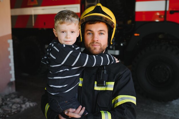 Dirty firefighter in uniform holding little saved boy standing on black background