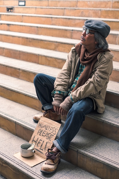 Dirty homeless person sit and looking on stairs with dollar cash in can