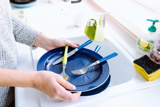 Грязные тарелки и столовые приборы в руках женщины, которая кладет их в кухонную раковину.
