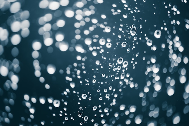 Dirty window glass with drops of rain. Atmospheric blue background with raindrops in bokeh. Droplets and stains close up. Detailed transparent texture in macro with copy space. Rainy weather.