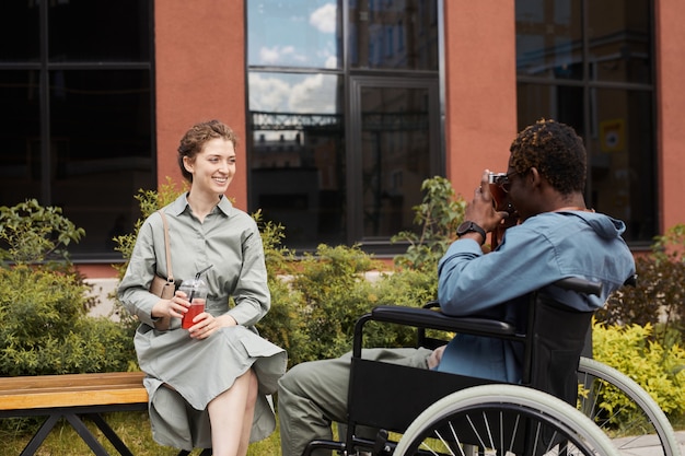 Disabled African-American photographer in wheelchair using camera while photographing young woman posing with summer beverage