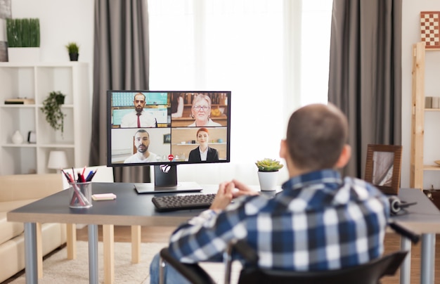 Uomo disabile in sedia a rotelle durante una videoconferenza con i colleghi di lavoro. giovane libero professionista immobilizzato che fa i suoi affari online, usando l'alta tecnologia, seduto nel suo appartamento, lavorando da remoto in