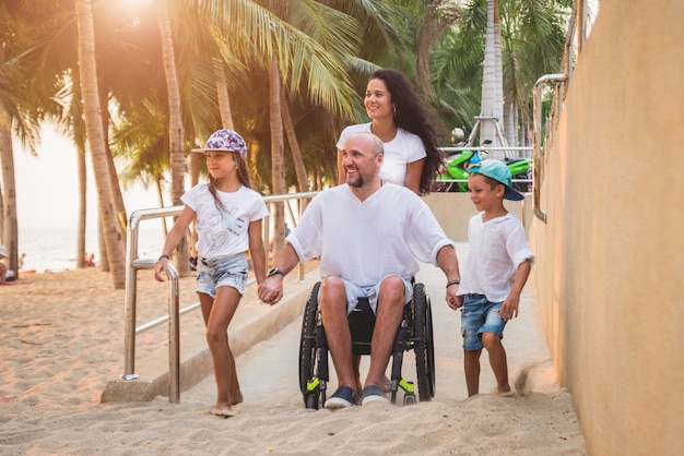 Disabled man in a wheelchair moves on a ramp to the beach with his family.