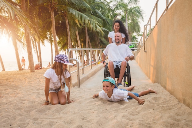 Disabled man in a wheelchair moves on a ramp to the beach with his family.