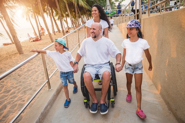 Disabled man in a wheelchair moves on a ramp to the beach with his family.