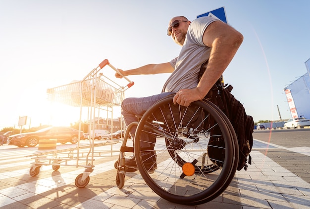 Инвалид в инвалидной коляске толкает тележку перед собой на парковке супермаркета