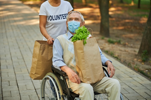 Vecchio disabile seduto sulla sedia a rotelle mentre tiene in mano una borsa della spesa di carta con del cibo