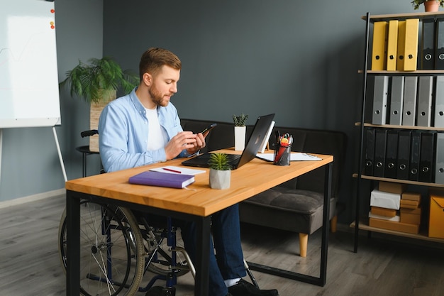 Disabled person in the wheelchair works in the office at the\
computer he is smiling and passionate about the workflow