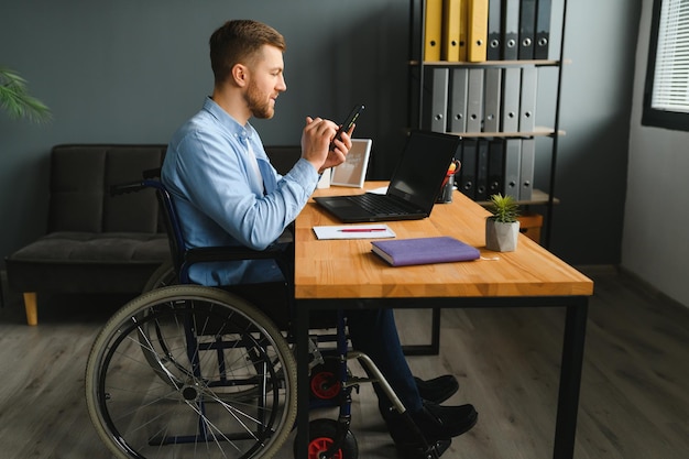 Disabled person in the wheelchair works in the office at the\
computer he is smiling and passionate about the workflow