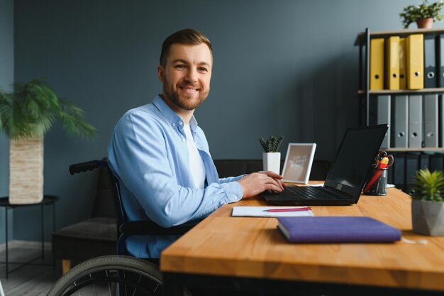 Disabled person in the wheelchair works in the office at the\
computer he is smiling and passionate about the workflow