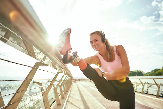 Disabled positive woman in sportswear and headphones stretching prosthetic leg while standing on the