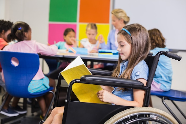 Foto ragazza disabile della scuola sulla sedia a rotelle che legge un libro in aula