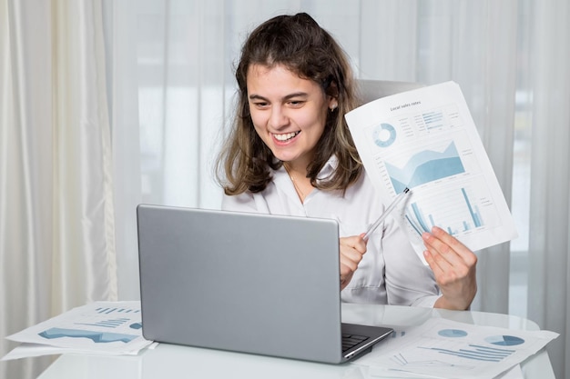 Disabled woman freelancer with infantile cerebral paralysis talks on videocall via laptop with manager showing project paper against window at home