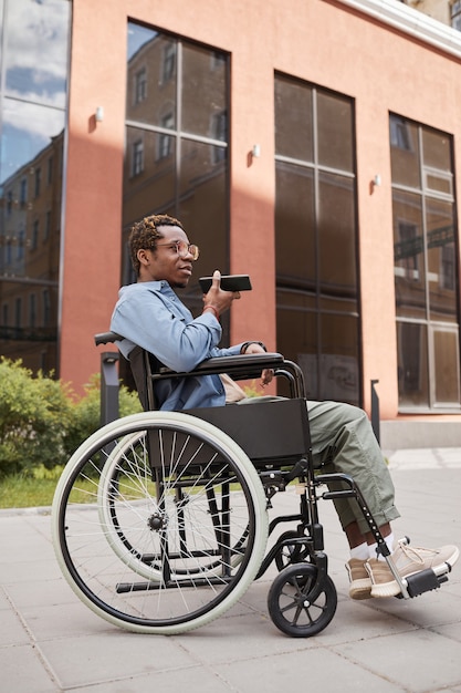 Молодой афроамериканец-инвалид в очках сидит в инвалидной коляске против современного здания и записывает голосовое сообщение на смартфон