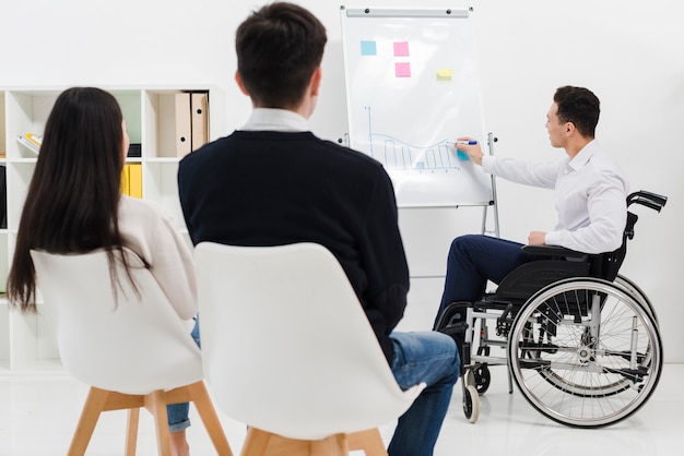 Foto giovane uomo d'affari disabile che si siede sulla sedia a rotelle che dà presentazione al suo collega di affari nell'ufficio