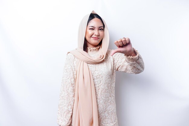 히잡을 쓴 실망한 아시아 무슬림 여성은 흰색 배경에 의해 격리된 반대의 손짓을 엄지손가락으로 내밉니다