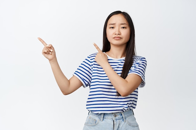 Disappointed asian woman frowning upset pointing fingers left at advertisement bad promotion express dislike and regret standing over white background