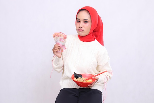 disappointed Asian woman wearing a hijab holding rupiah notes and a bowl containing ramen Chinese fo