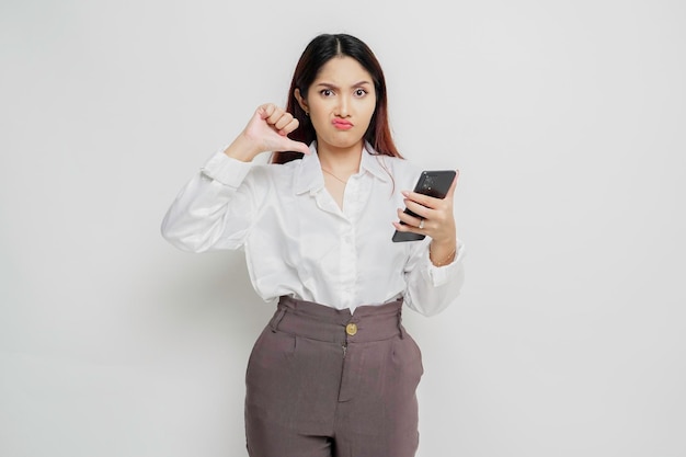 A disappointed Asian woman wearing white shirt gives thumbs down hand gesture of disapproval isolated by white background