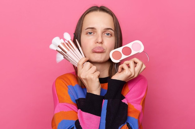 Disappointed displeased woman wearing sweater holding cosmetic brush powder looking with pout lips being in bad mood posing isolated on pink background