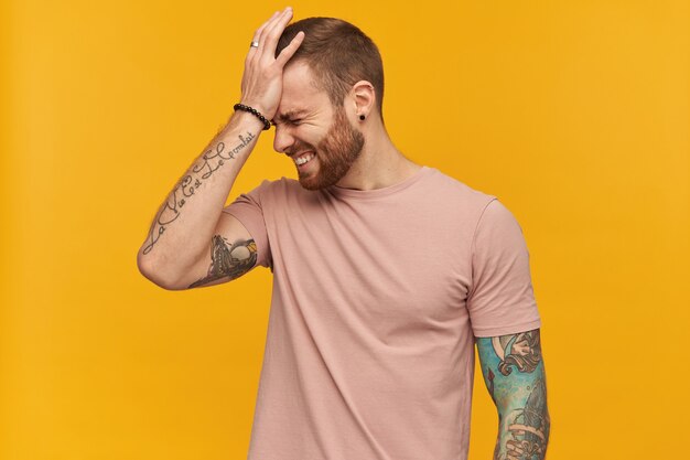 Disappointed embarrassed young bearded man with tattoo and eyes closed in pink tshirt keeps hand on forehead and looks upset over yellow wall