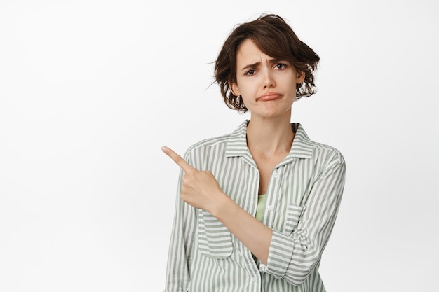 Disappointed frowning girl, sulking upset, pointing finger at upper left corner and furrow eyebrows skeptical, standing on white