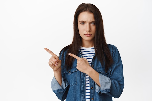 Disappointed frowning woman pointing left, looking serious and reluctant, dislike promotion, so low discount, waiting for explanation, standing upset against white background