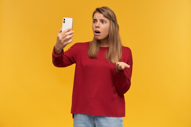 Disappointed girl keeps mobile phone in her hand, looking on it like making a video call, opened her mouth like being misunderstood