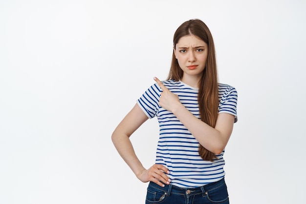 Disappointed girl pointing left frowning and looking upset at camera standing with skeptical face expression against white background